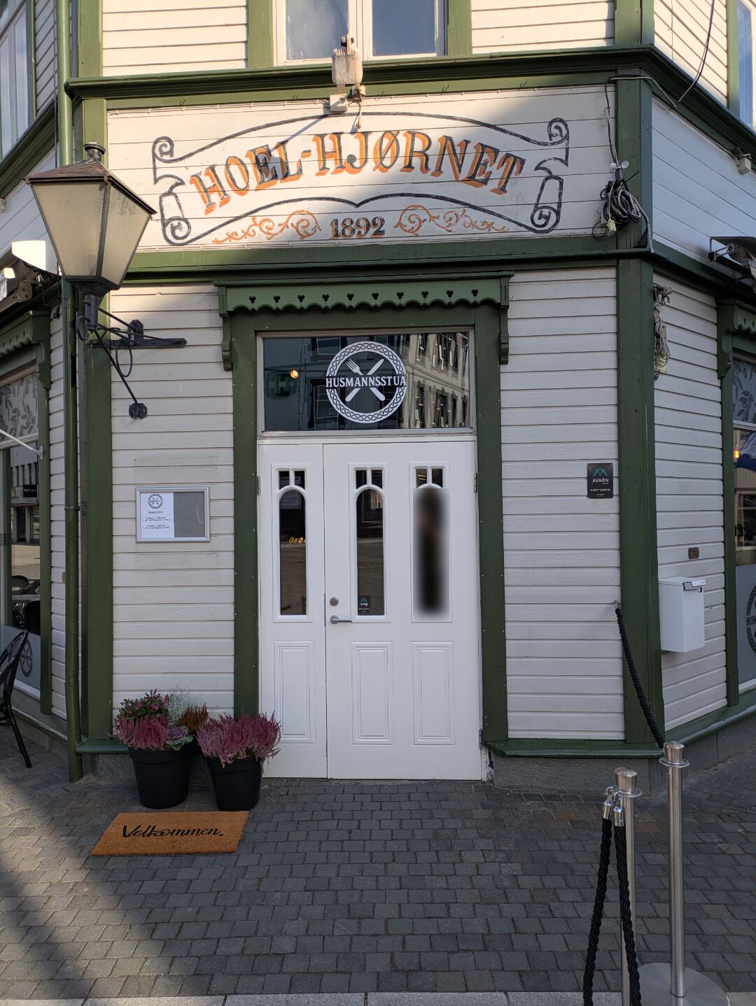 Inngangen til Husmannsstua på hjørnet: hvit dobbeltdør med logoen i glasset, skiltet «Hoel-Hjørnet 1892» over døra og en velkommen-matte foran